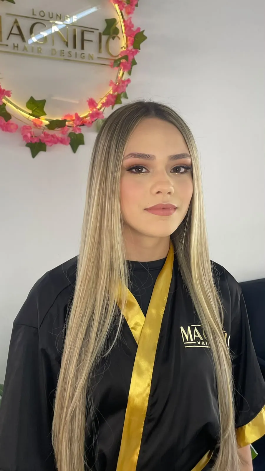 Loiro longo e liso com raiz esfumada, cliente de frente ao letreiro do Lounge Magnific